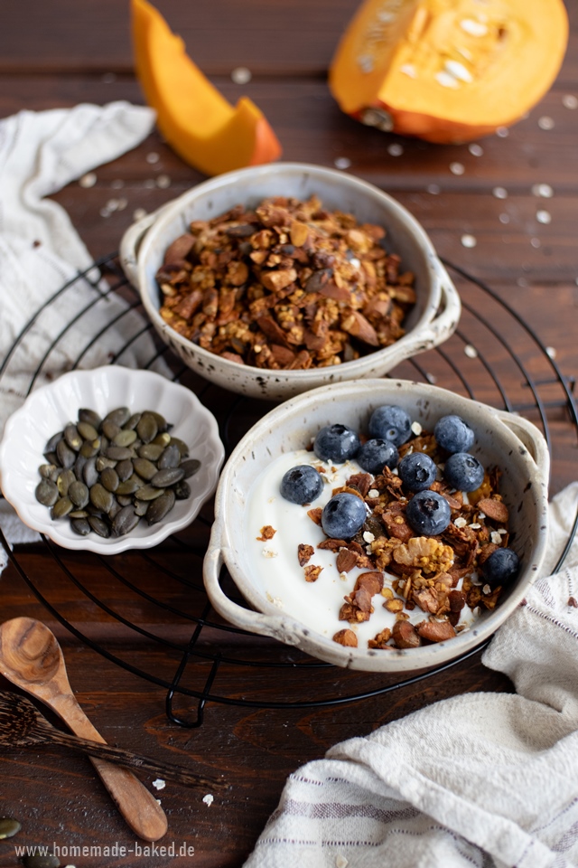 Zwei Schalen mit selbstgemachtem Crunchy-Kürbis-Granola stehen auf einem runden Gitter. Eine Schale enthält das Granola pur, die andere ist mit Joghurt und frischen Blaubeeren angerichtet. Daneben stehen eine kleine Schale mit Kürbiskernen und Holzlöffel. Im Hintergrund sind Kürbisstücke und Haferflocken auf einem rustikalen Holztisch zu sehen.