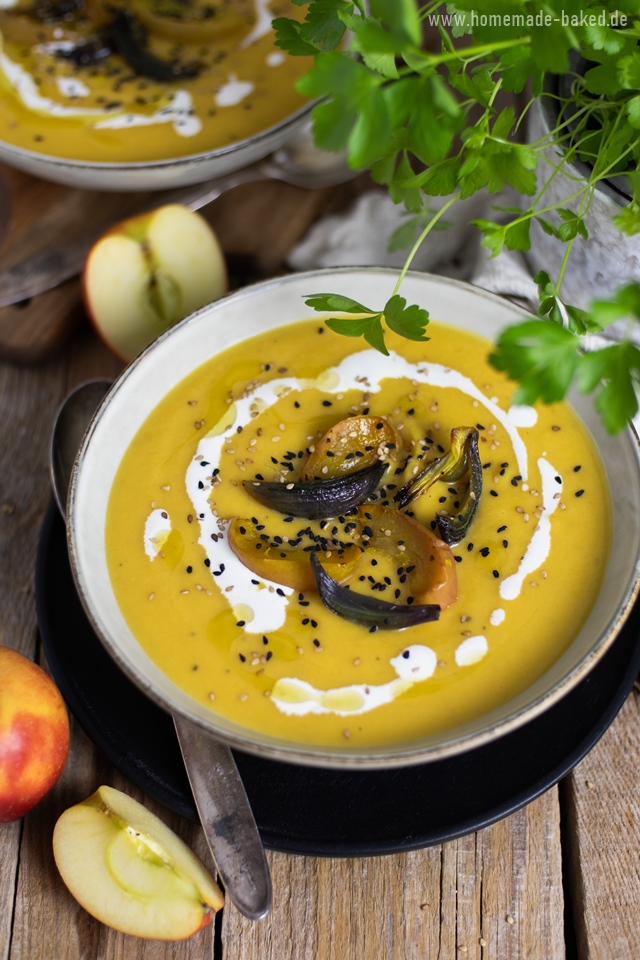 Eine ofengeröstete Kürbissuppe in einer Schüssel, garniert mit gerösteten Apfel- und Zwiebelscheiben, Schwarzkümmel, Sesam und einem Klecks Sahne. Im Hintergrund sind frische Äpfel und grüne Kräuter zu sehen. Die Suppe wird auf einem rustikalen Holztisch serviert.